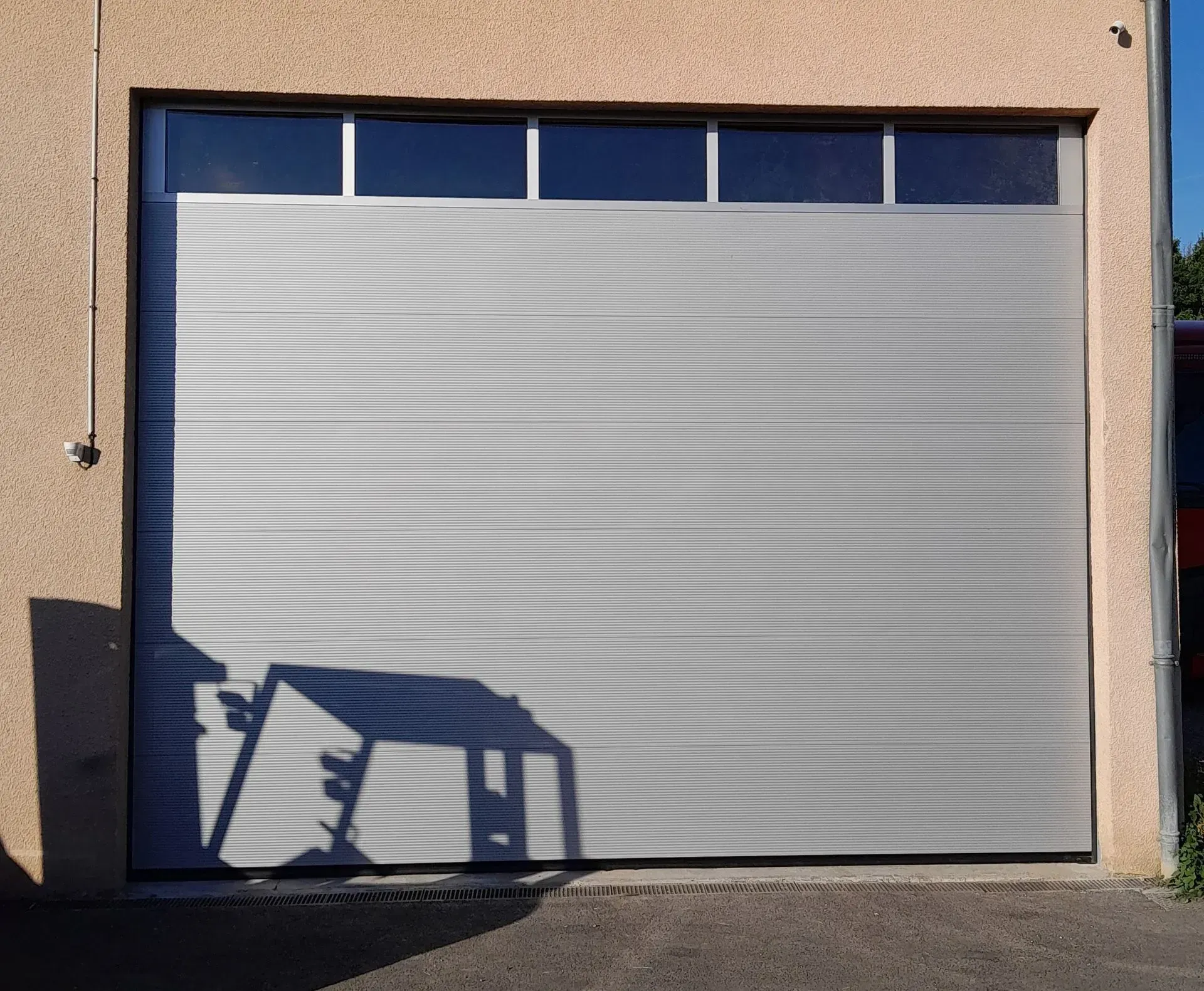 Porte de garage sectionnelle argentée fermée sur un bâtiment beige aux finitions bleues, avec l'ombre d'une chaise devant.