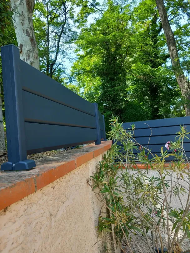 Clôture en bois bleu foncé le long d'un mur beige, avec des arbres verts en arrière-plan.