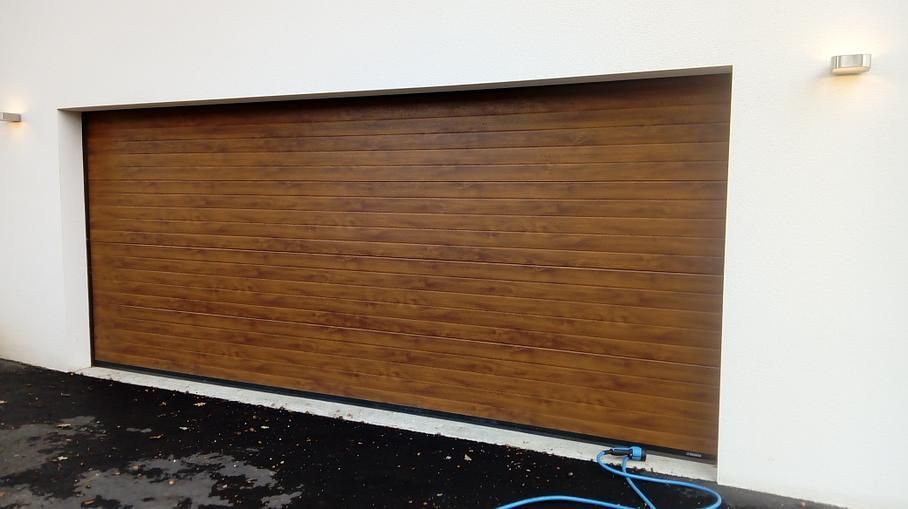 Porte de garage en bois brun fermée sur un mur extérieur blanc, avec un trottoir mouillé en dessous.