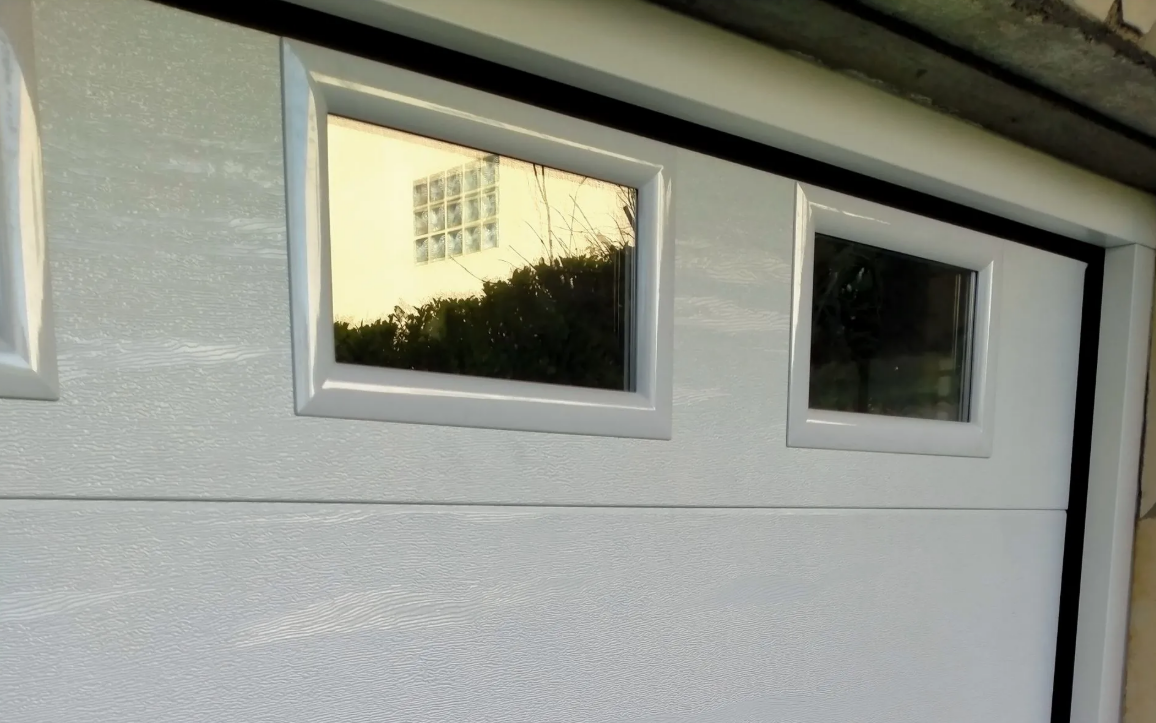 Porte de garage blanche avec deux petites fenêtres reflétant des arbres et un bâtiment