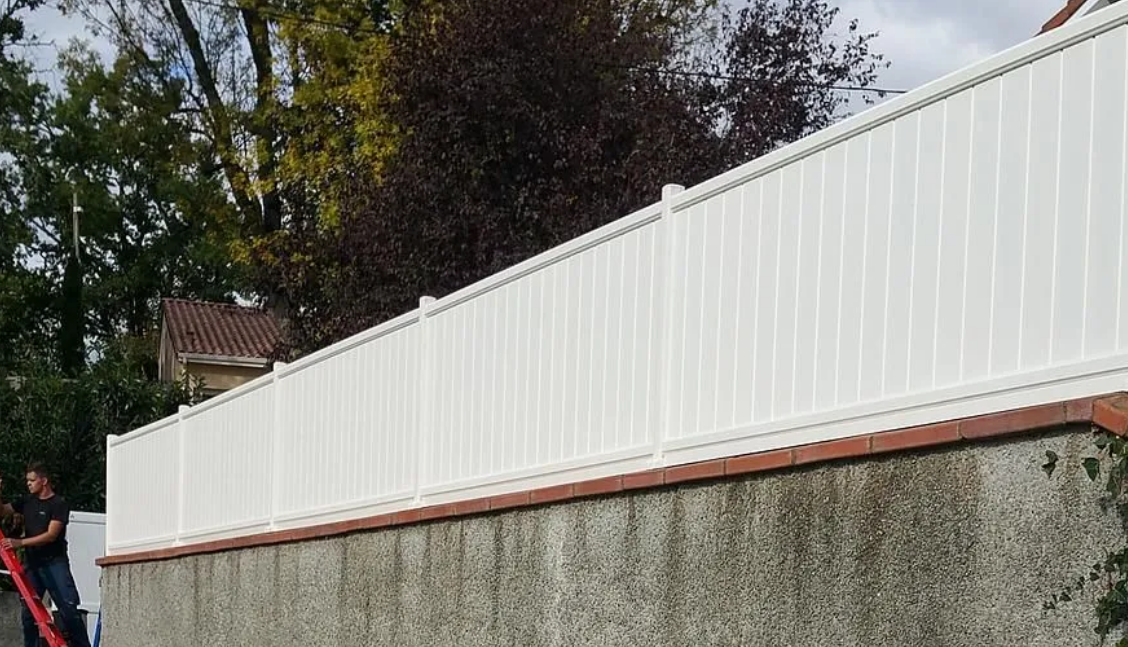 Clôture blanche préservant l'intimité, surmontant un mur en béton, avec des arbres et des toits visibles derrière.
