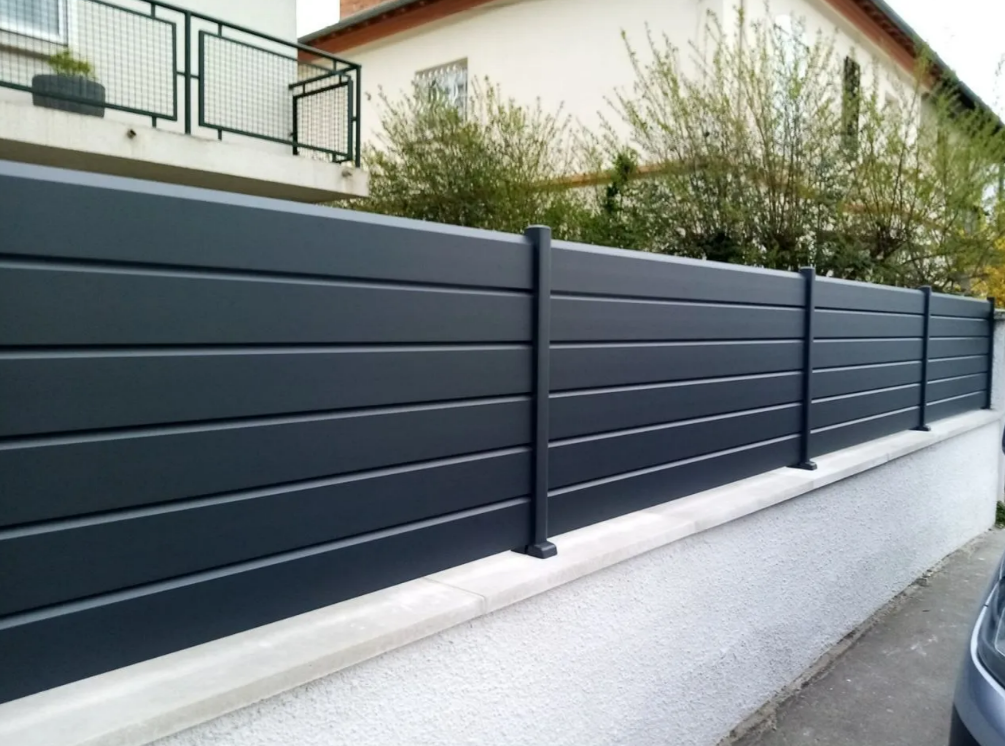 Clôture à claire-voie gris foncé le long d'un mur de soutènement blanc, à côté d'une maison et d'arbustes.