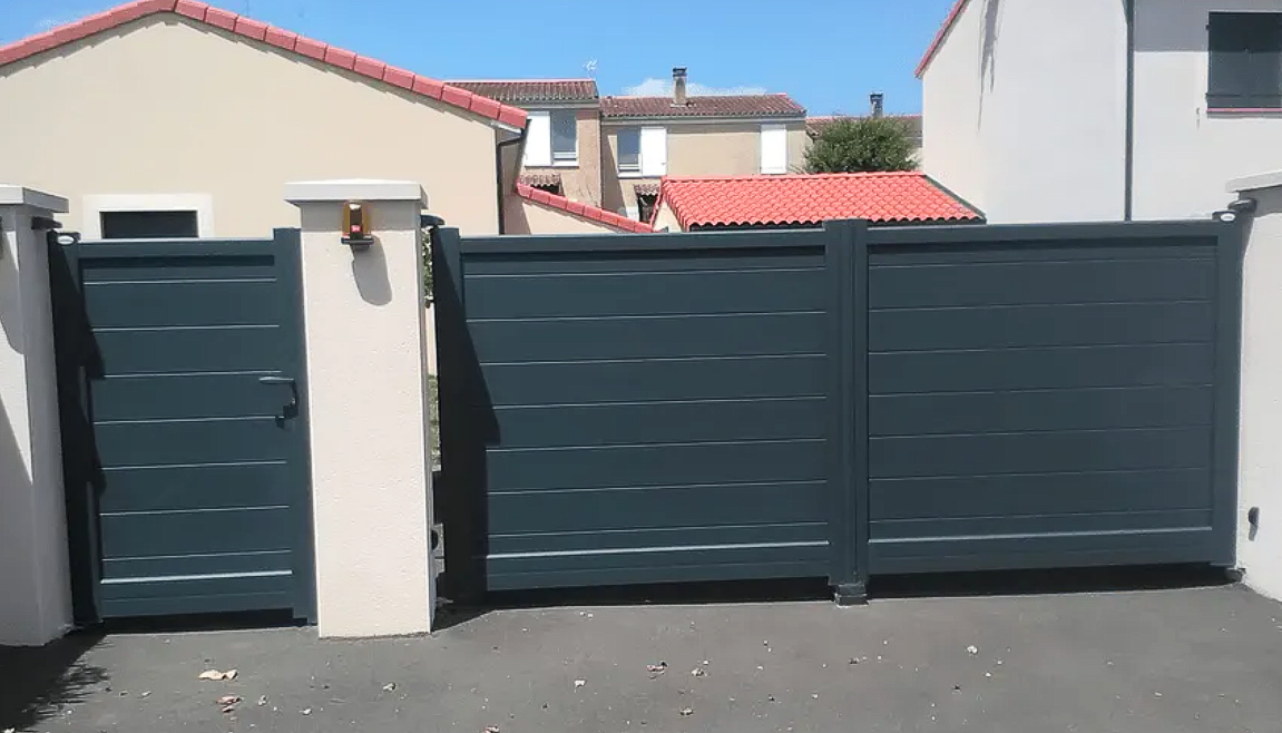 Portail d'entrée et porte latérale en métal gris, jouxtant un mur blanc dans une cour résidentielle.