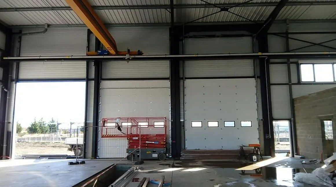 Intérieur d'un grand entrepôt industriel avec poutres en acier, portes de garage et une nacelle élévatrice rouge près du centre.