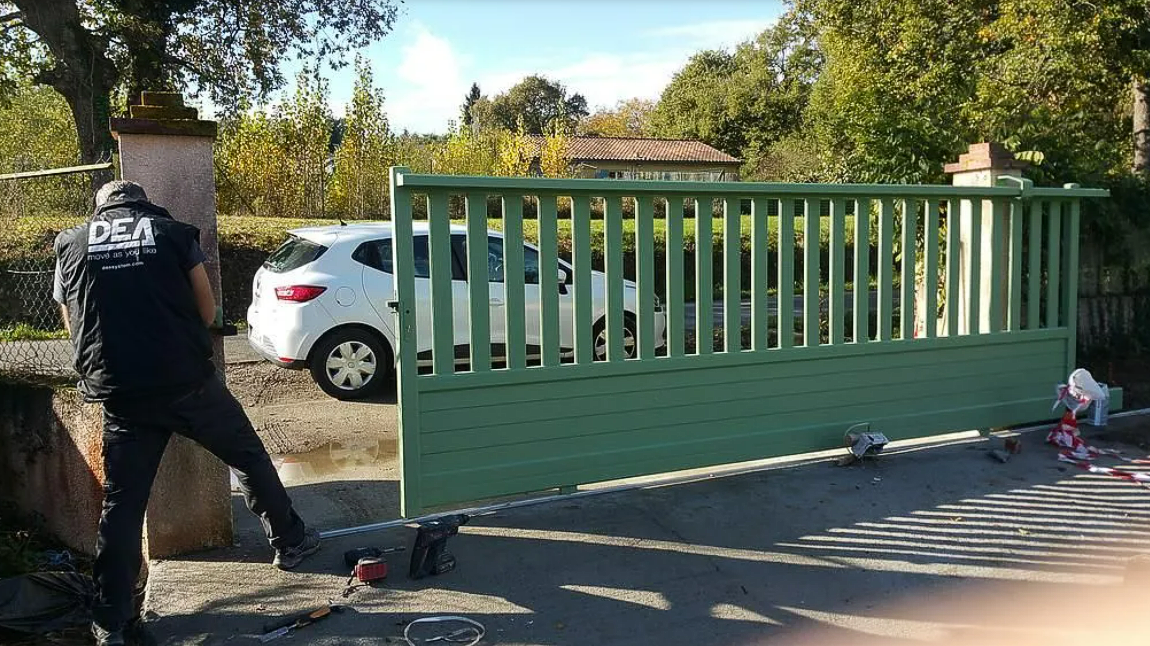 Une personne passe devant un portail métallique vert à côté d'une voiture garée dans une rue ensoleillée.
