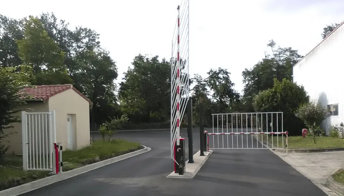 Portail à l'entrée d'une allée avec barrières rouges et blanches, arbres et petit bâtiment à proximité