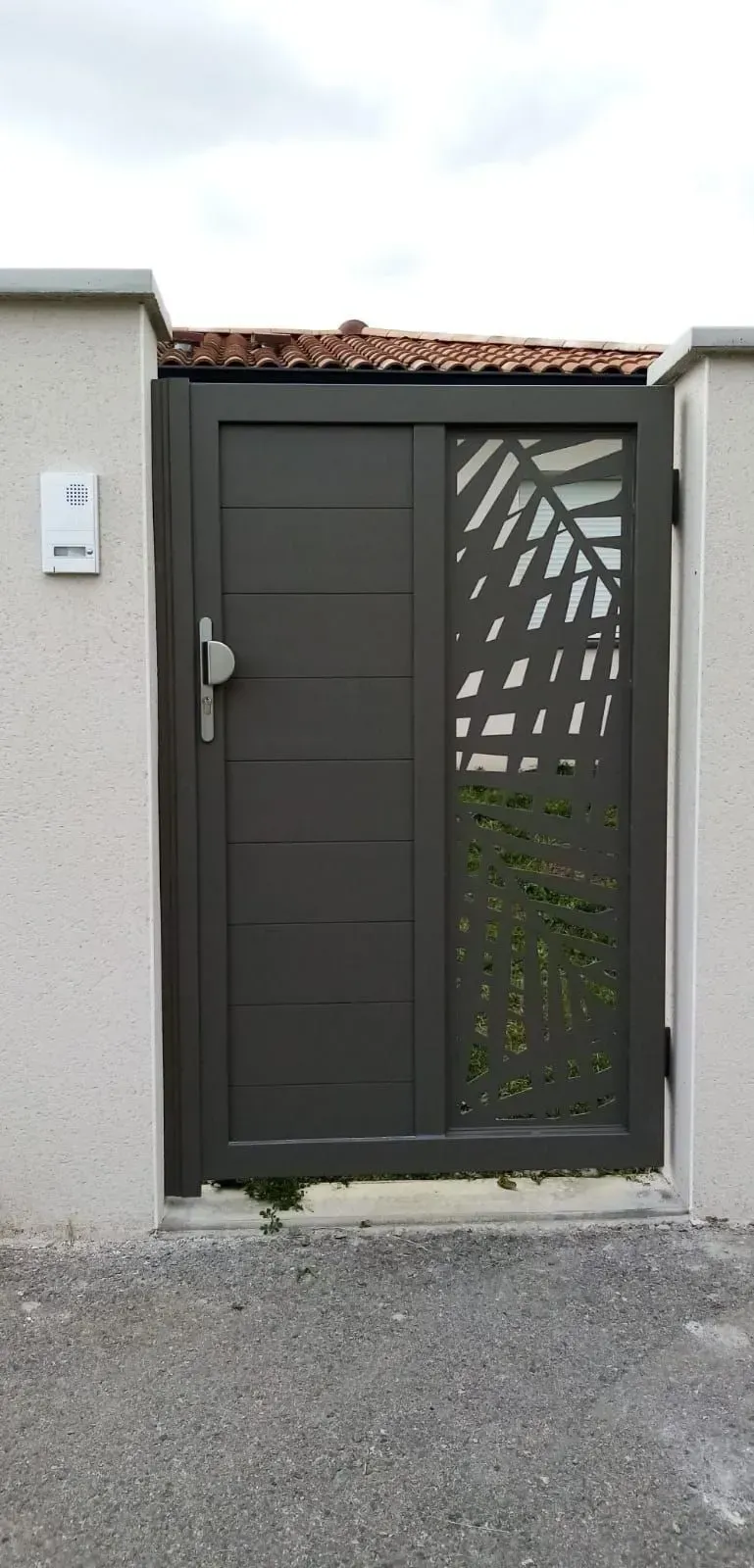 Portail en métal gris foncé avec porte piétonne et panneau décoratif en treillis dans un mur en béton.