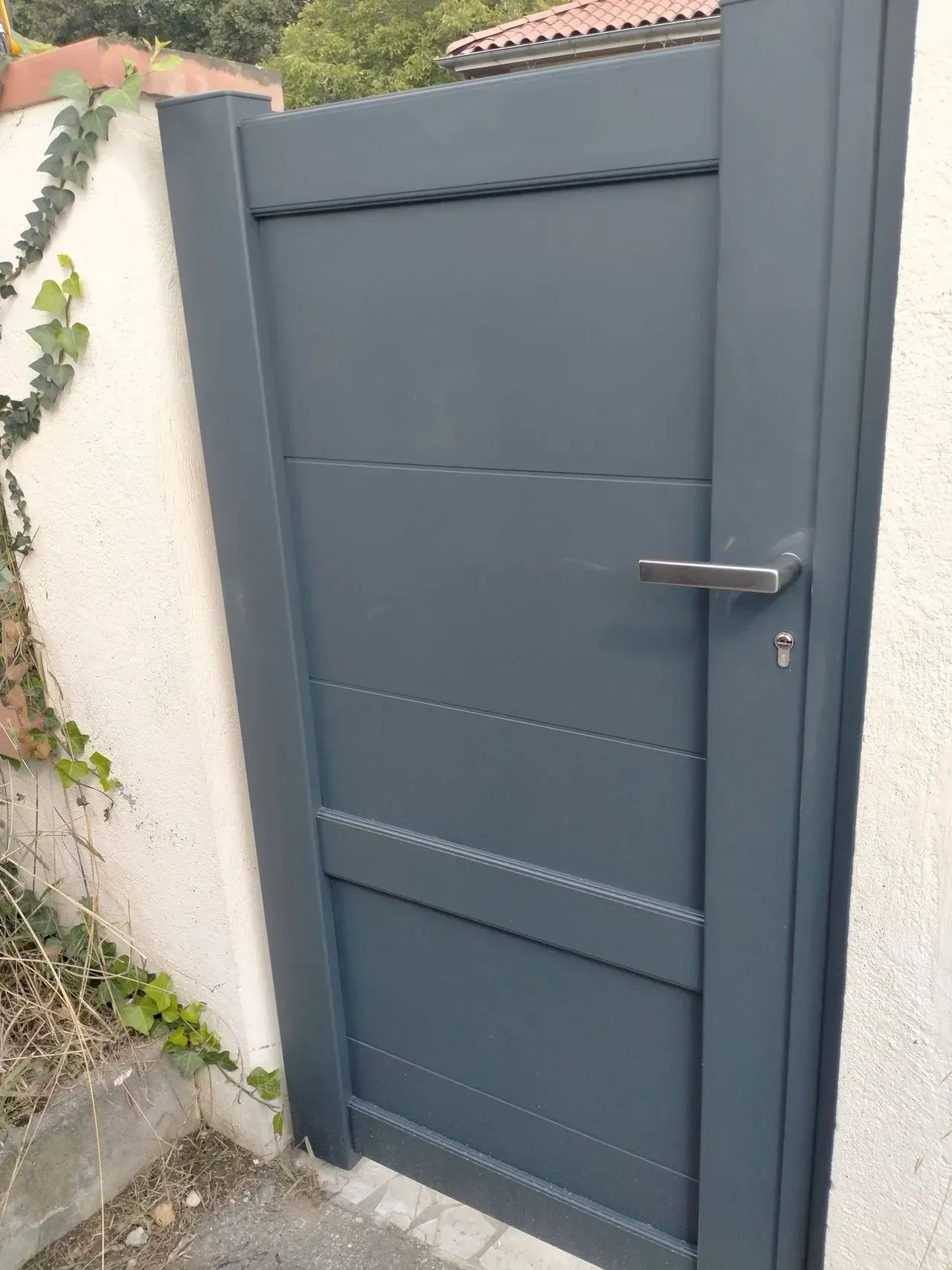 Portail en métal gris avec poignée et serrure encastrés dans un mur extérieur en stuc blanc.