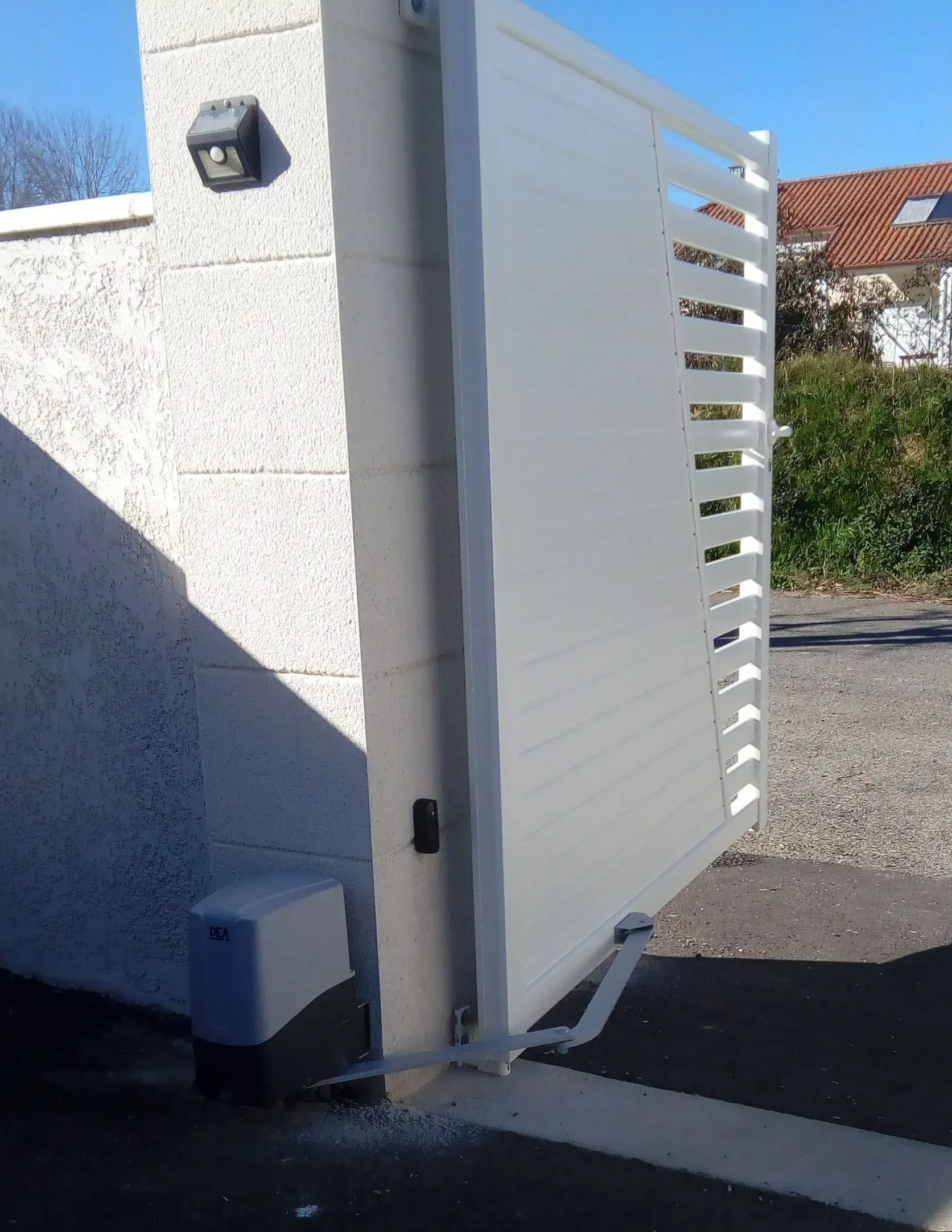 Portail blanc à claire-voie, adossé à un mur en stuc, partiellement ouvert sur une allée baignée de soleil.