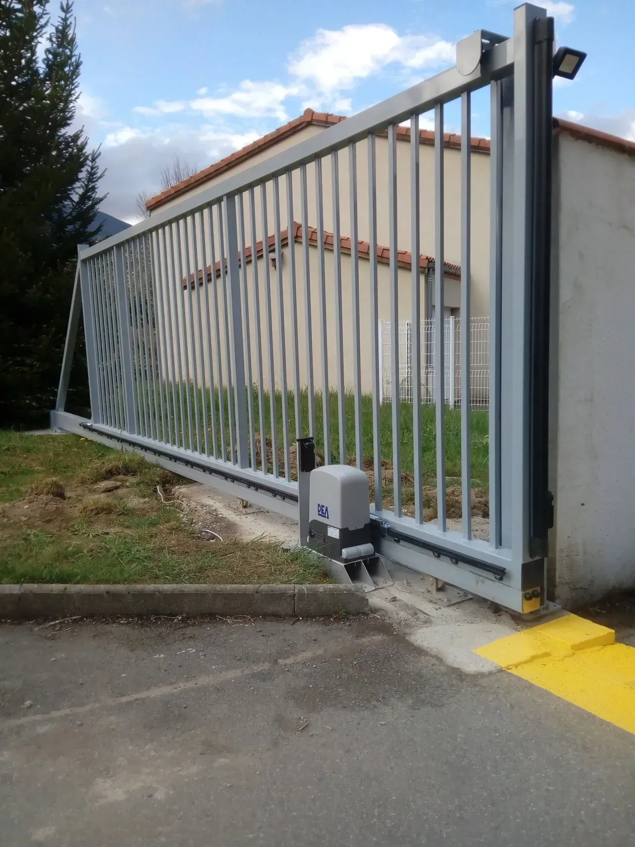 Portail coulissant métallique barrant une allée carrossable à côté d'un bâtiment, avec un petit moteur de portail à sa base.