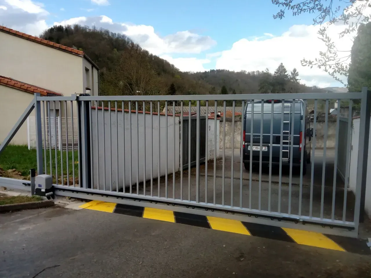 Portail coulissant en métal barrant une allée bordée d'un trottoir noir et jaune, avec une camionnette garée et une colline en arrière-plan.