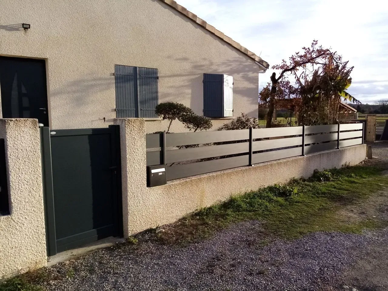Maison avec un mur en stuc beige, un portail vert foncé et une clôture basse le long d'une allée de gravier.