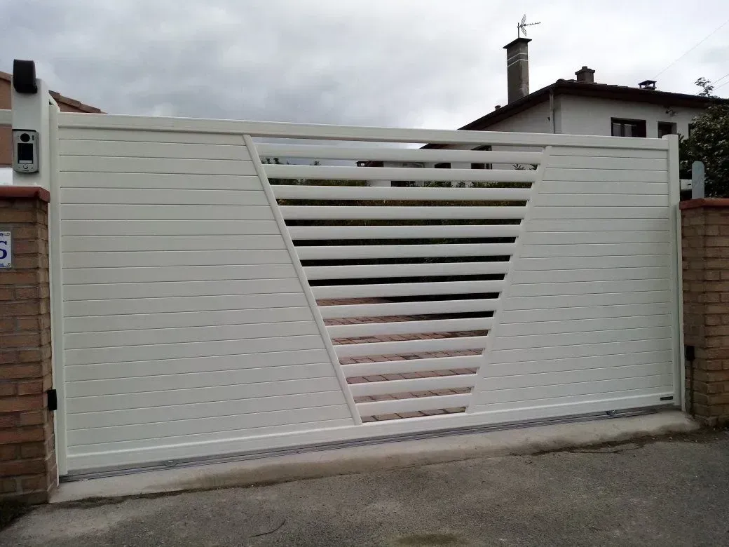 Portail coulissant blanc avec lames diagonales et piliers en briques sous un ciel nuageux