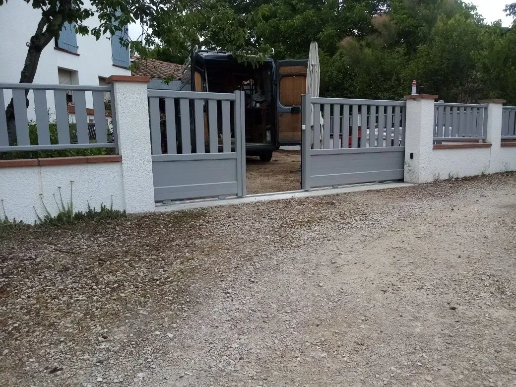 Portail en métal gris dans une clôture blanche le long d'une allée de gravier, avec des arbres et une maison en arrière-plan.