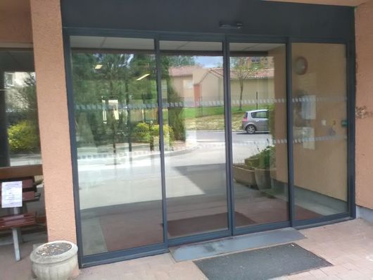 Entrée vitrée avec cadres sombres, petit paillasson et reflets de la rue.