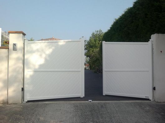 Portail d'allée blanc à deux panneaux fermés donnant sur une entrée pavée, arbres visibles derrière.