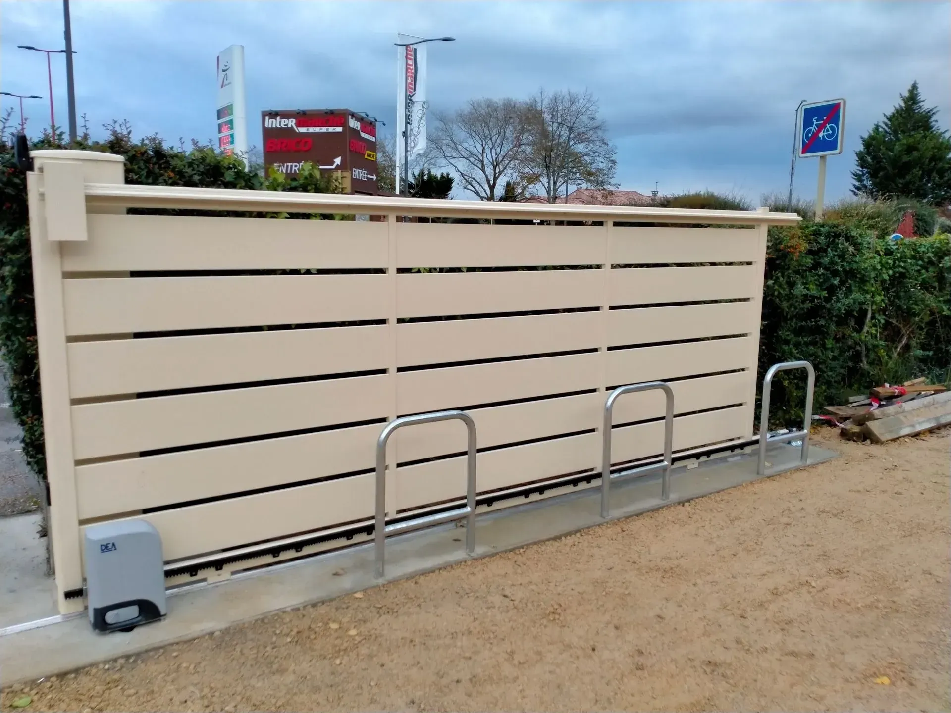Un abri utilitaire beige à claire-voie jouxte un trottoir, avec des supports à vélos en métal et des haies en arrière-plan.