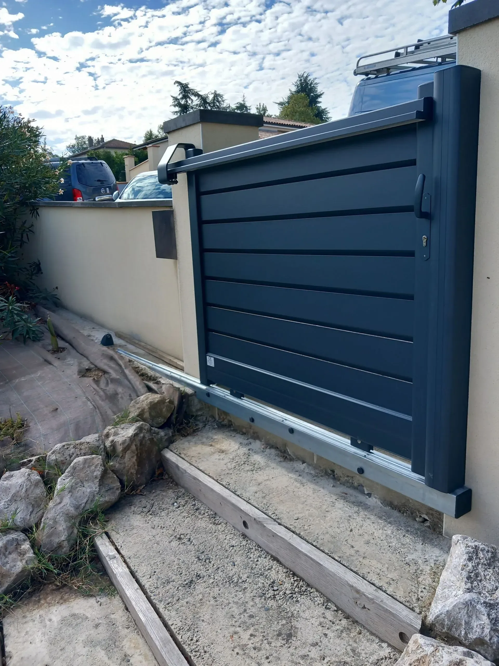 Portail coulissant bleu au bout d'une allée en béton, à côté d'un mur beige et d'un chemin rocailleux.