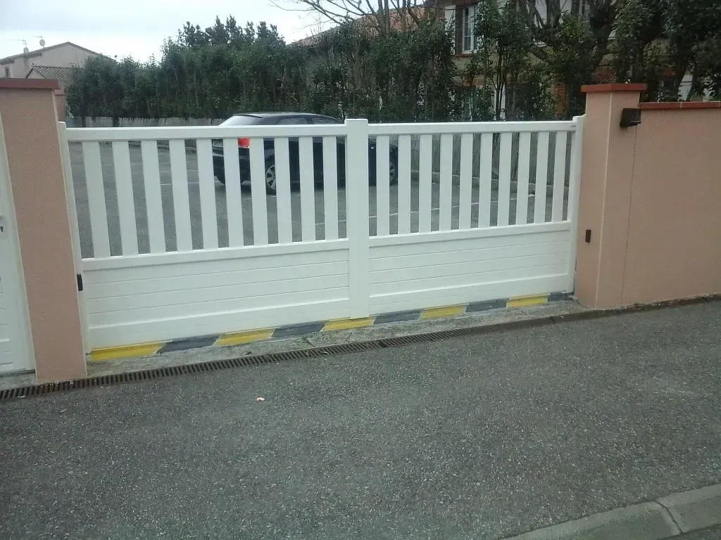 Portail coulissant blanc entre des murs couleur pêche, au bord d'une route pavée.