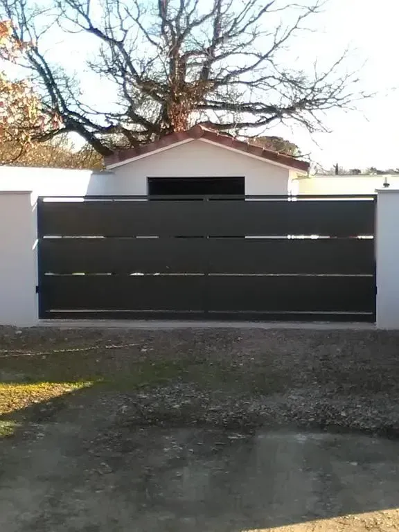 Portail coulissant noir devant un garage blanc et arbre sans feuilles dans une allée.