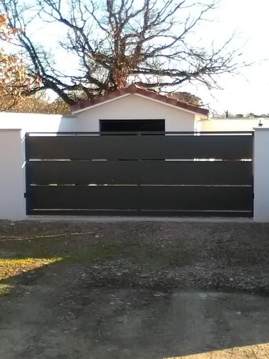Portail en métal noir devant un garage ou un abri de jardin blanc, avec un arbre au-dessus et une allée de gravier.
