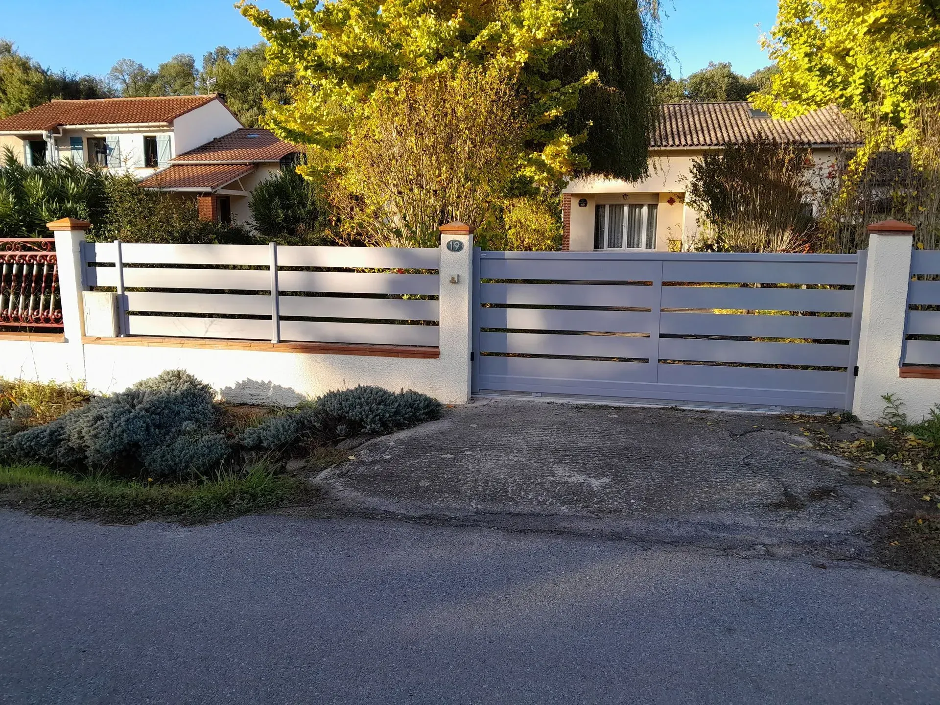 Portail et clôture blancs à claire-voie devant une maison, avec des arbres et des arbustes derrière.