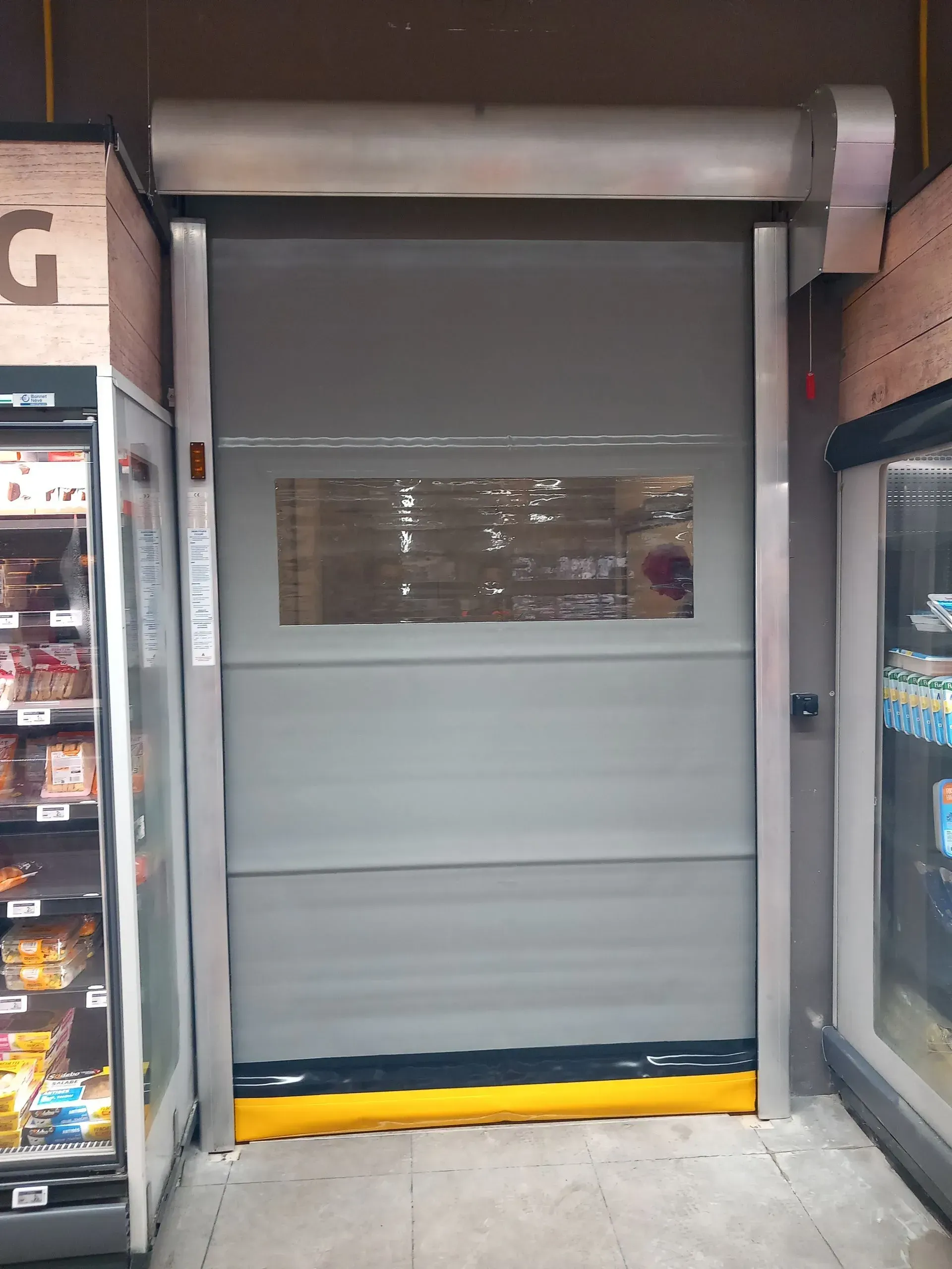 Volet de sécurité gris à enroulement recouvrant l'entrée d'un magasin, avec une bande de protection jaune en bas.