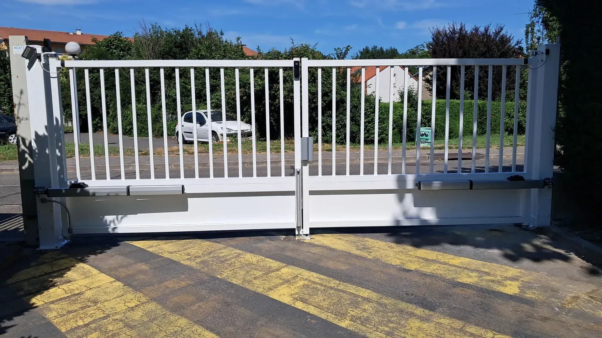 Portail coulissant en métal blanc barrant une allée donnant sur une rue et des maisons, marquages ​​routiers jaunes devant.