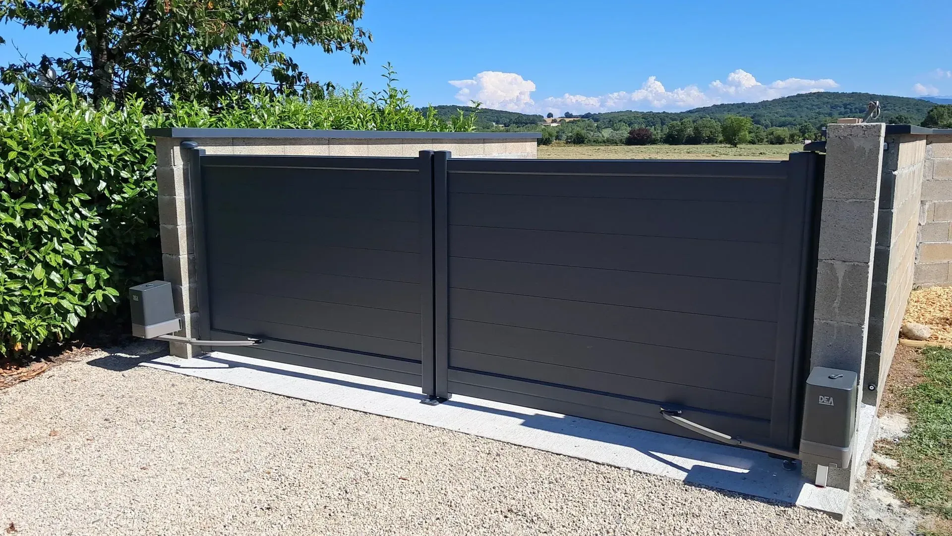Portail coulissant en métal sombre à côté d'un pilier en pierre sur une allée de gravier sous un ciel bleu