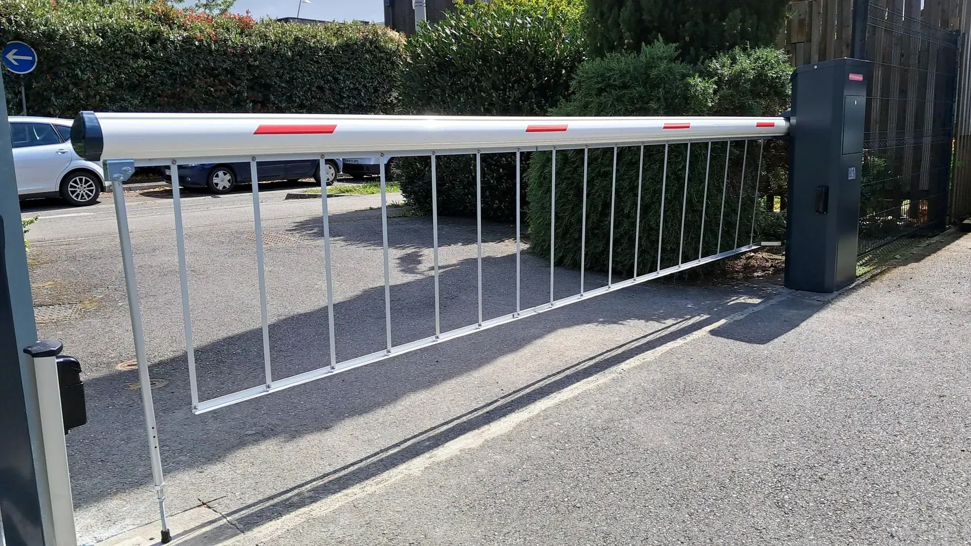 Barrière de parking blanche abaissée en travers d'une allée longeant une route goudronnée et un terrain gravillonné.