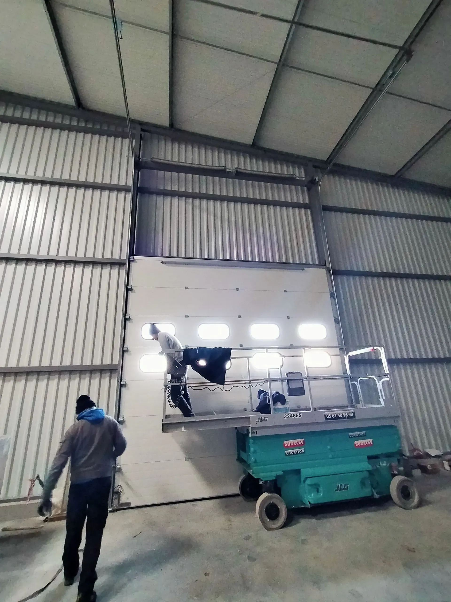 Un ouvrier se tient près d'une nacelle élévatrice verte dans un entrepôt dont la grande porte industrielle est ouverte et l'intérieur baigné de lumière.