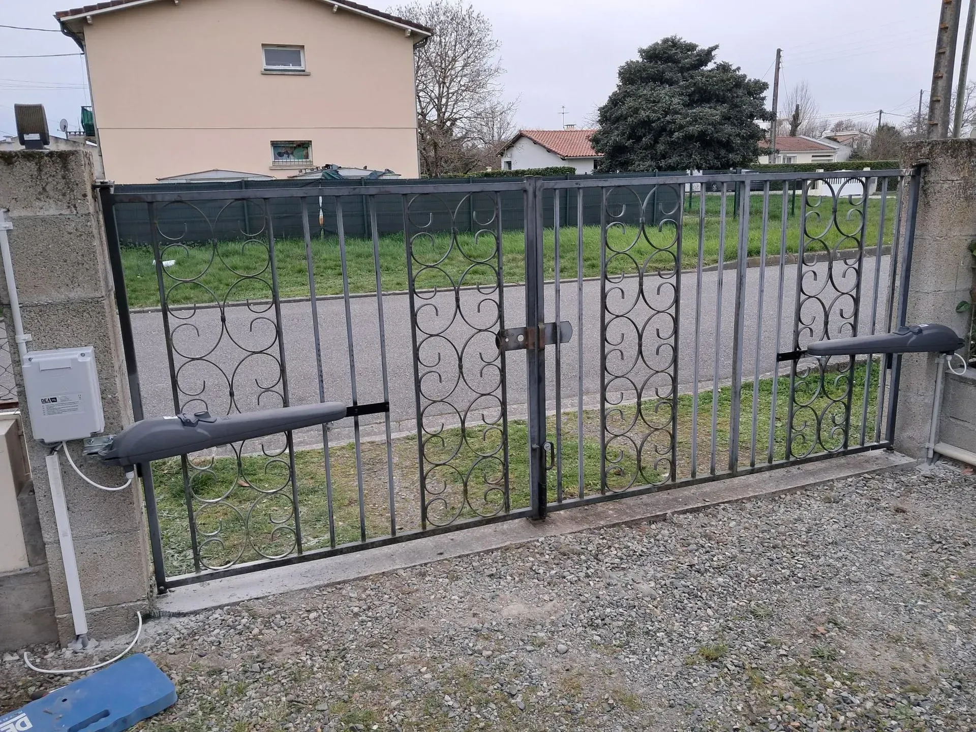 Un portail métallique noir fermé barre l'allée de gravier devant une maison et un jardin roses.