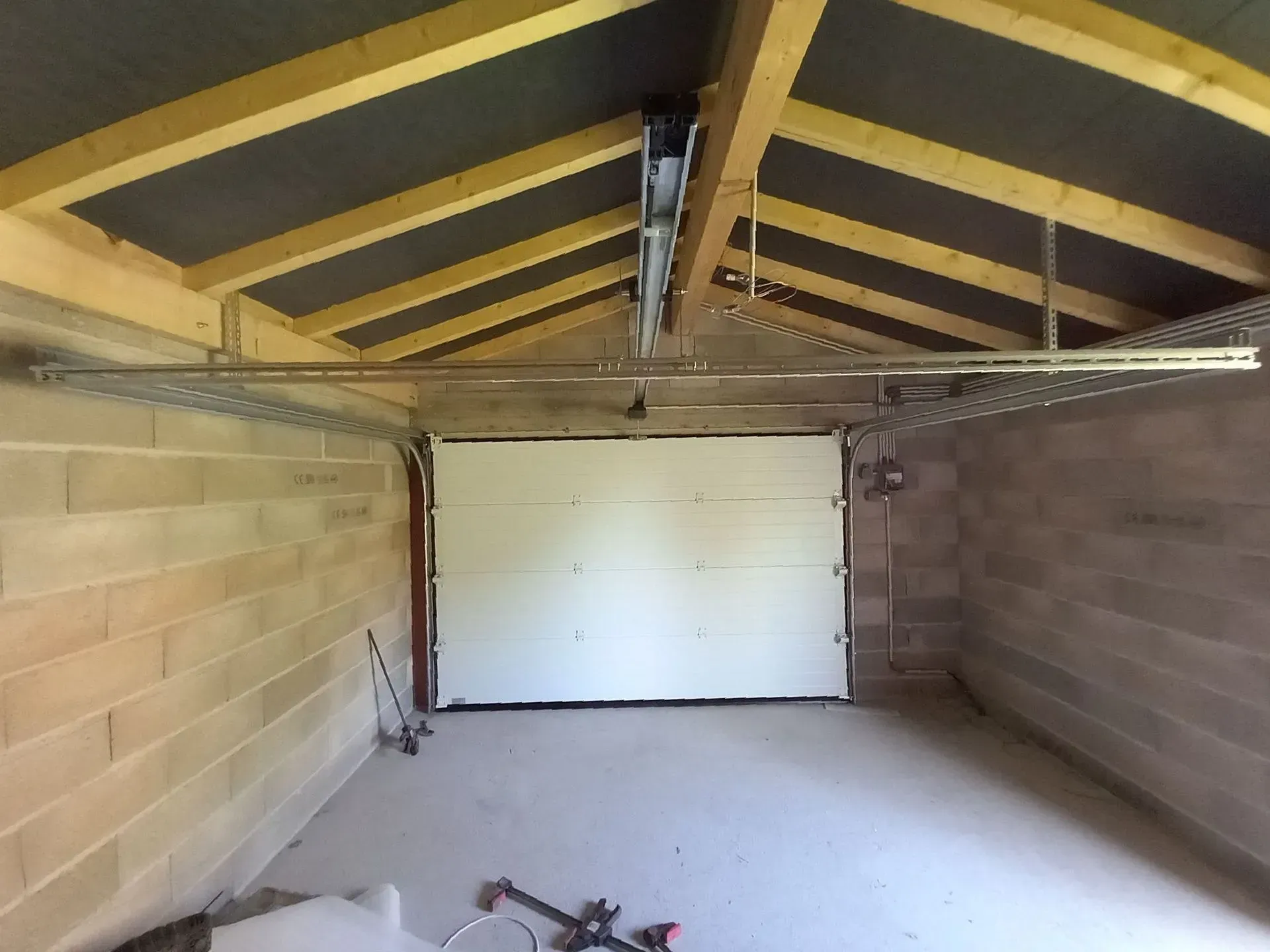 Garage vide avec une porte blanche, des poutres apparentes au plafond et un sol en béton brut.