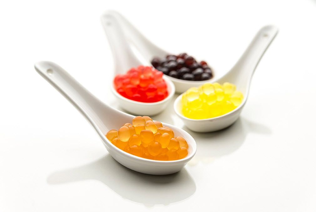 Drei Löffel gefüllt mit verschiedenfarbigen Geleekugeln auf einem weißen Untergrund.