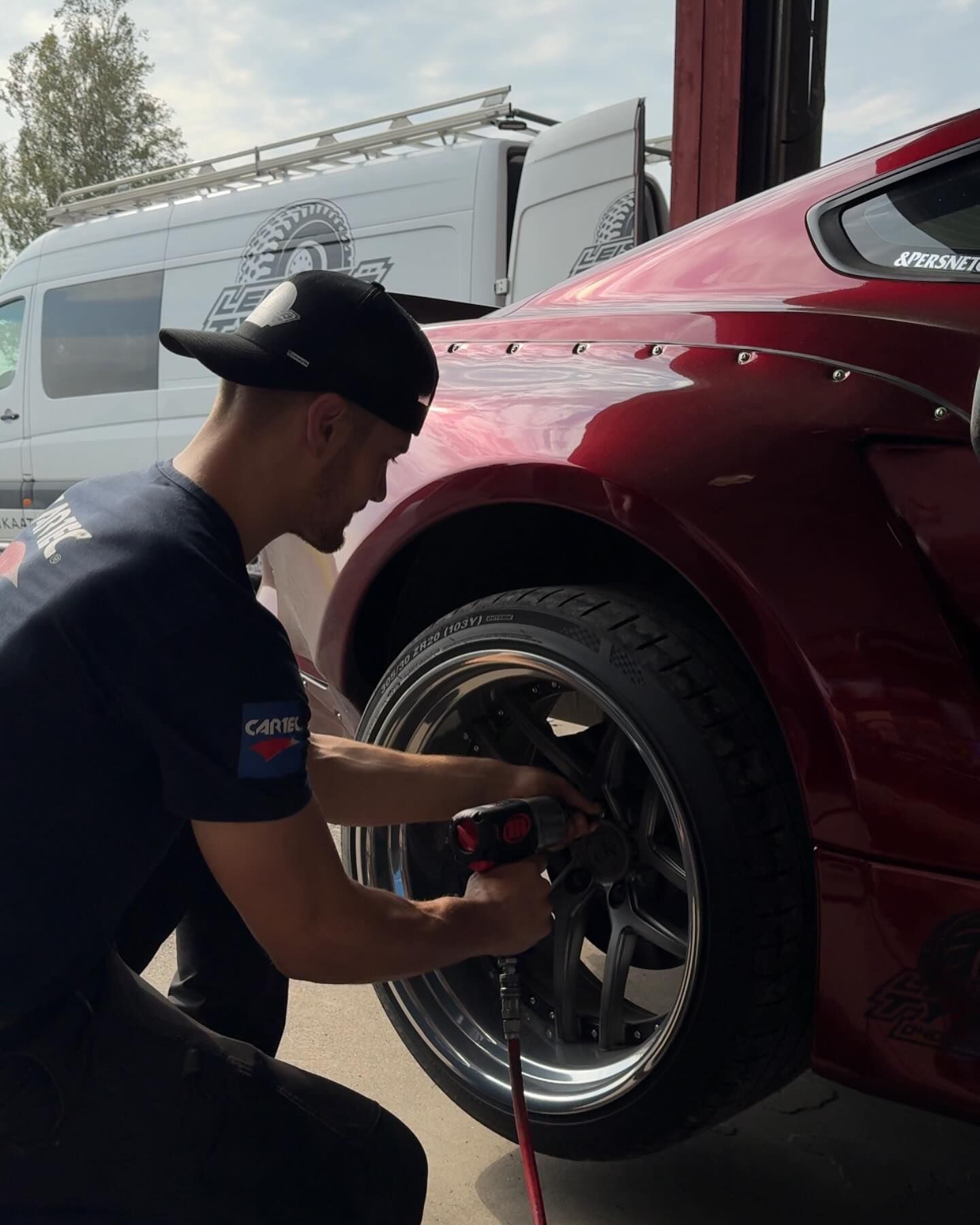 Mekaanikko käyttää sähkötyökalua punaisen auton vanteella ulkona pakettiauton lähellä.
