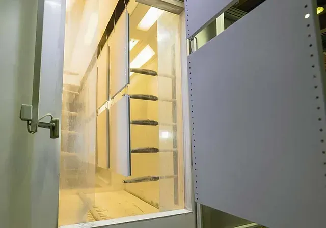 Varios paneles blancos rectangulares cuelgan en el interior de una cabina de pintura industrial con luces brillantes en el techo.