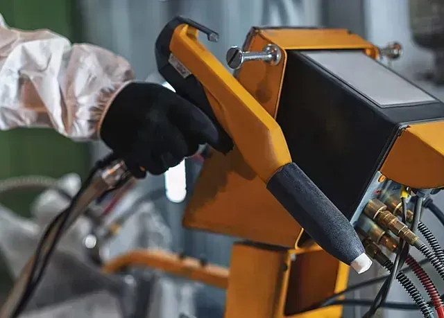 Una persona que lleva un traje protector blanco y guantes negros utiliza una pistola pulverizadora de pintura en polvo industrial de color amarillo.