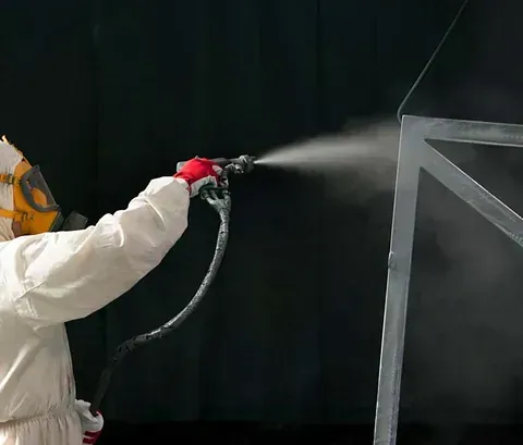 Una persona con un traje protector y una mascarilla respiratoria rocía pintura sobre una estructura metálica sobre un fondo oscuro.