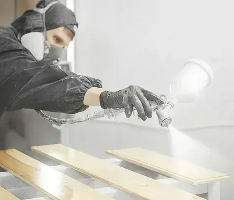 Un trabajador con equipo de protección utiliza una pistola pulverizadora para aplicar un acabado blanco a tablas de madera en un taller.