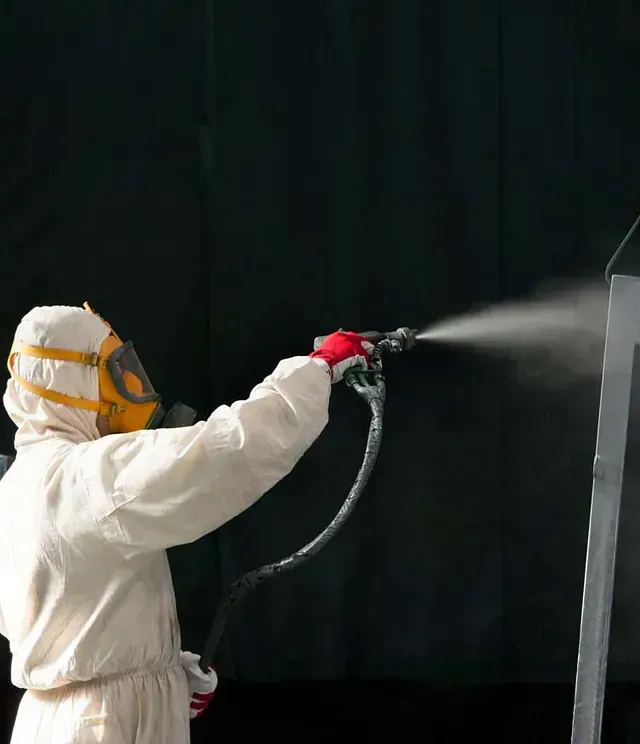 Una persona con traje protector y máscara respiratoria utiliza una pistola pulverizadora para aplicar un acabado a una superficie sobre un fondo oscuro.