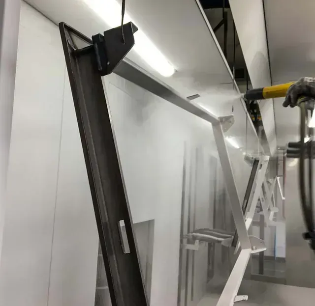 Una pistola pulverizadora aplica una capa de pintura en polvo blanca a una estructura metálica que cuelga en una cabina de pintura industrial.
