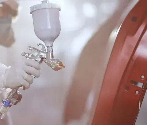Una persona con guantes blancos utiliza una pistola pulverizadora para aplicar una capa de pintura roja a un panel de un coche.