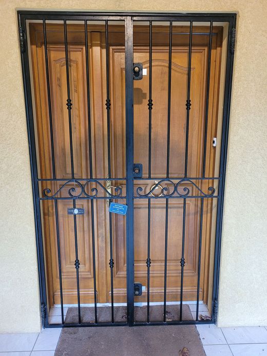 Une double porte en bois est sécurisée par une grille de sécurité en fer forgé ornée de volutes décoratives et d'une serrure.