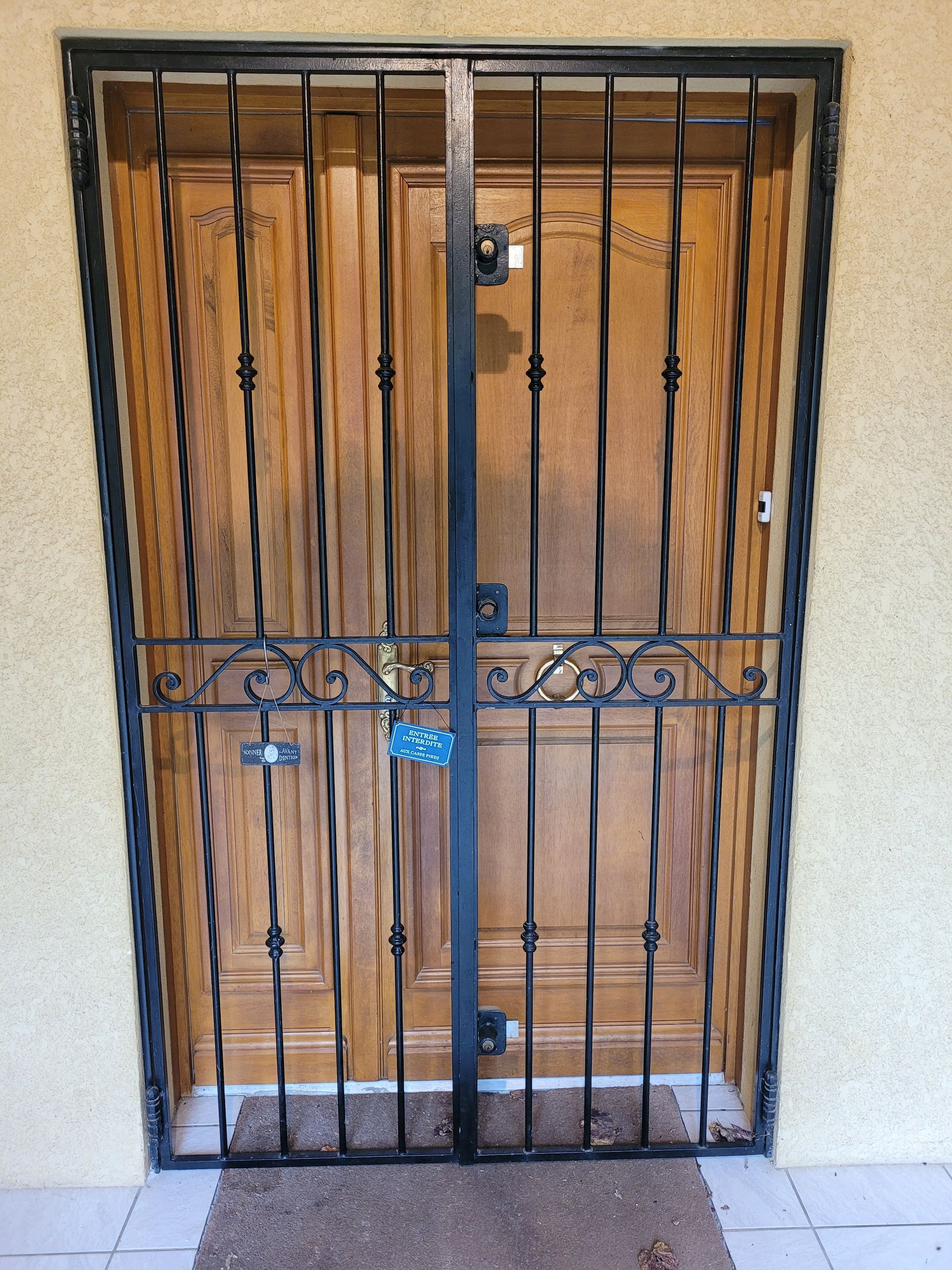 Une double porte en bois sécurisée par une grille de sécurité en métal ornée de volutes horizontales et d'un cadenas.
