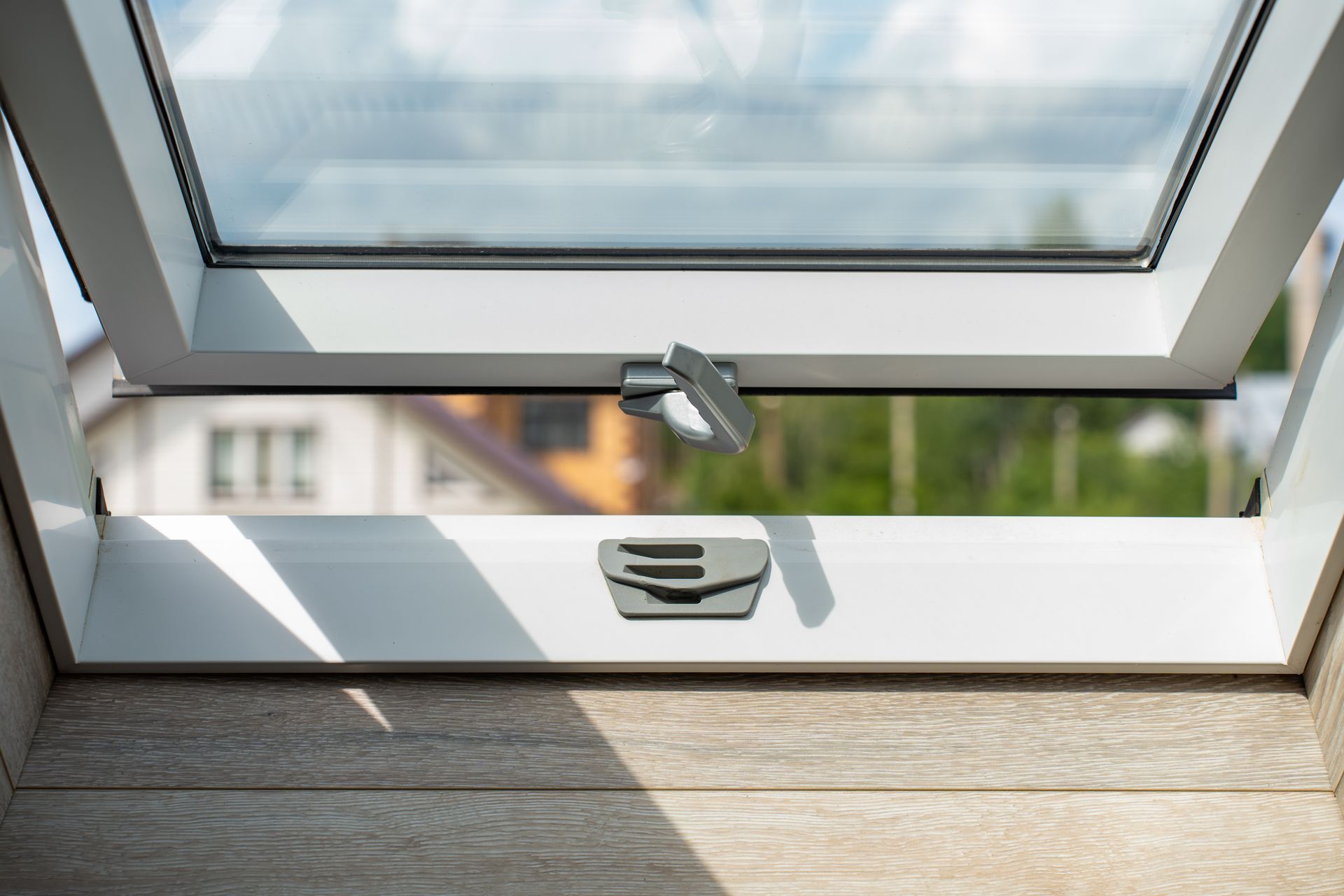 Une fenêtre de toit blanche est partiellement ouverte, donnant sur un arrière-plan flou de maisons et d'arbres.