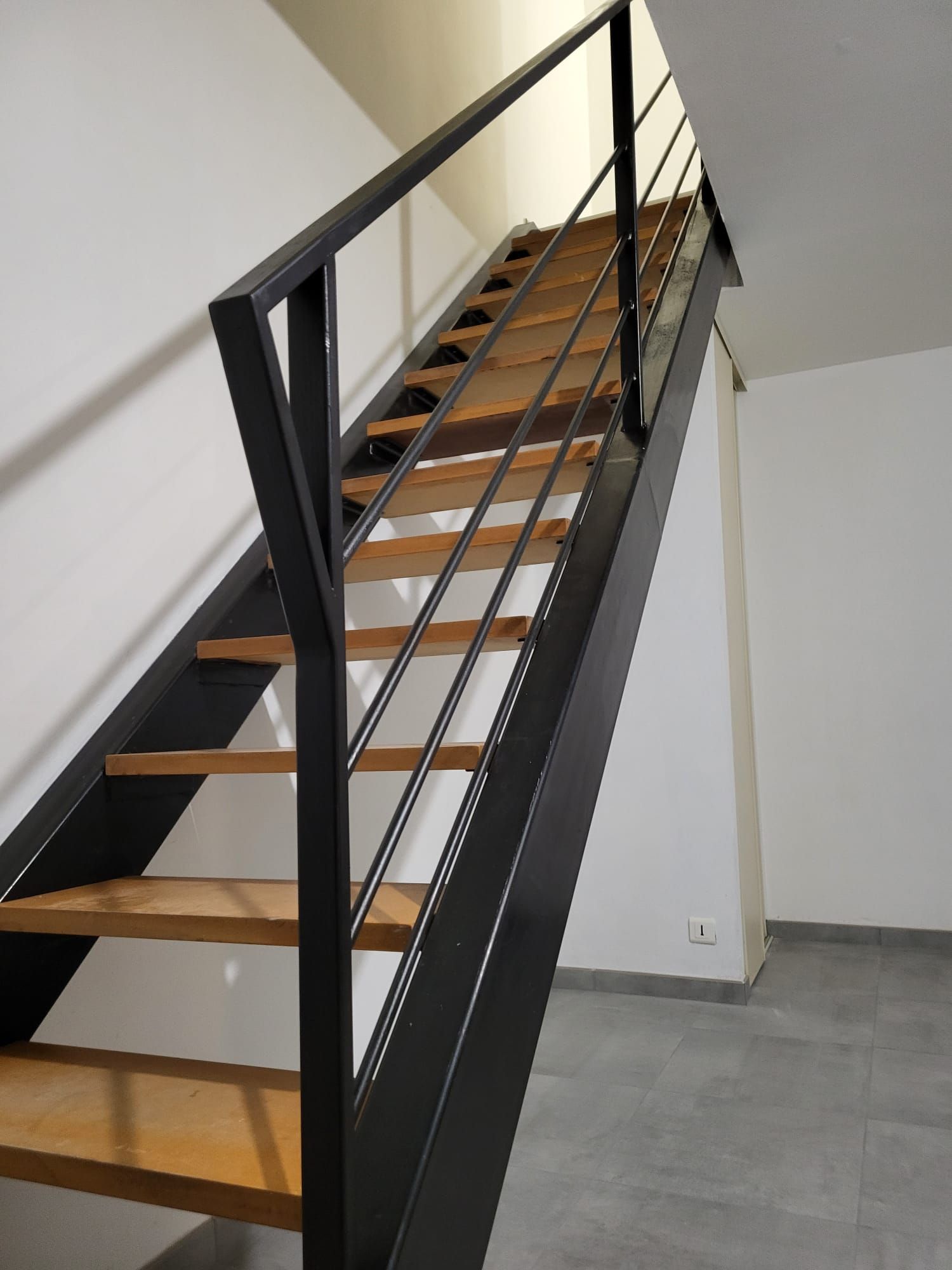 Un escalier courbe avec une rampe en métal sombre ornée se détache sur un mur texturé dans un couloir intérieur.