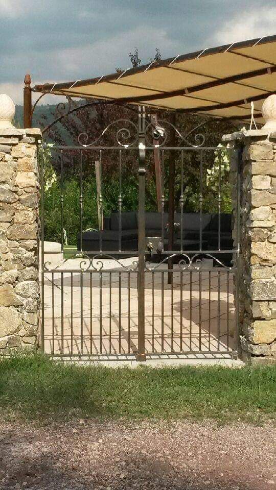 Un portail en métal sombre, encadré par deux piliers de pierre, s'ouvre sur un patio ombragé par un auvent.