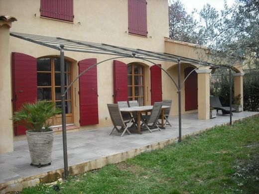 Un patio avec une pergola en métal, une table, des chaises et une plante en pot devant une maison aux volets et à la terrasse en pierre.