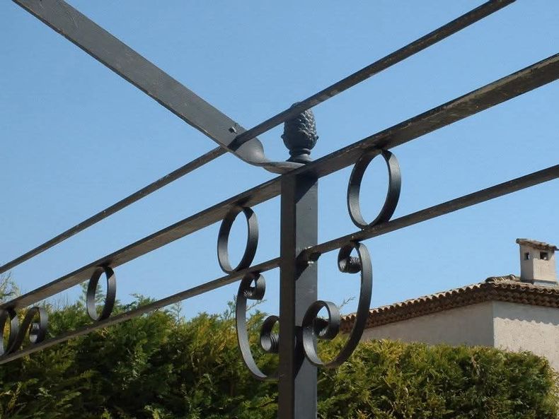 Détail d'angle d'une pergola en fer, orné d'éléments décoratifs circulaires et de volutes.