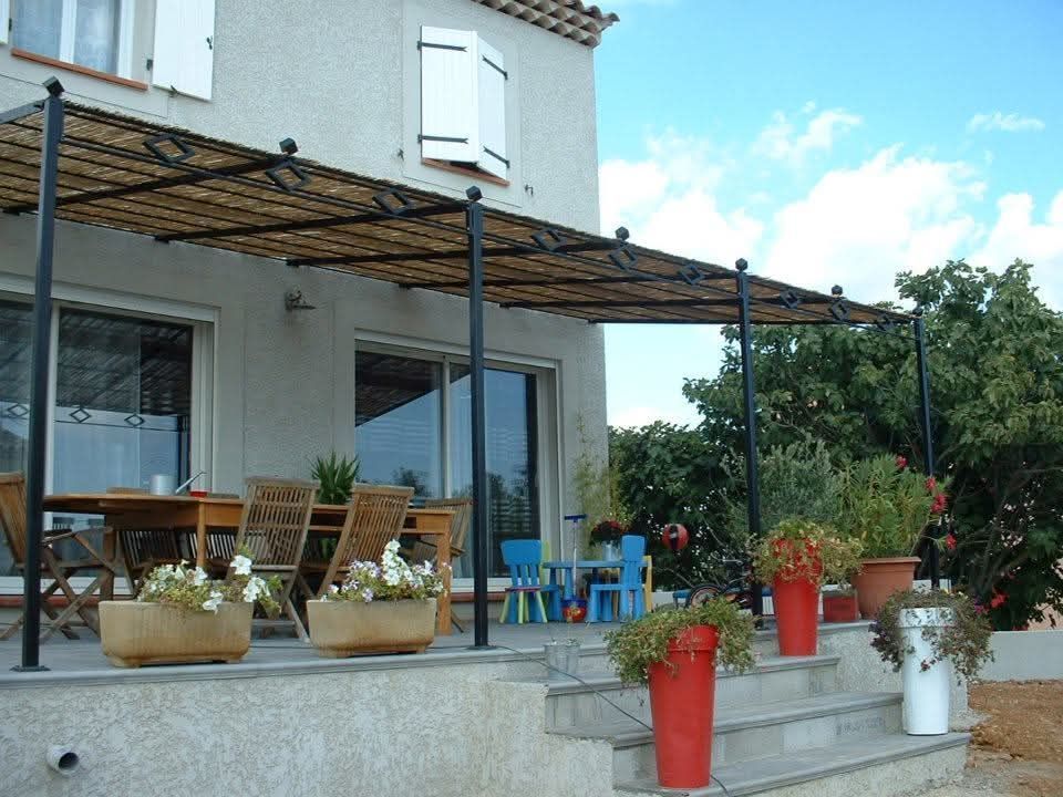Un patio couvert d'une treille en bambou, avec du mobilier de jardin et des plantes en pot, aménagé sur une terrasse en pierre près d'une maison.